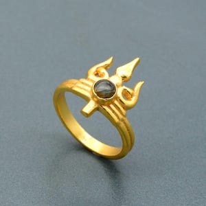 Puede incluir: Anillo dorado con diseño de tridente y una piedra oscura en el centro. El anillo tiene una apariencia detallada y ornamentada, con las púas del tridente y la punta central claramente definidas. El anillo está sobre un fondo gris sólido.