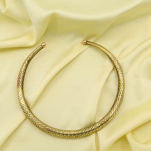 May include: A gold-colored collar necklace with a textured, scale-like pattern. The necklace has a rounded shape and small ball-shaped ends. The jewelry is displayed on a light yellow satin fabric.