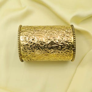 May include: A gold-colored cuff bracelet with an intricate floral pattern. The bracelet has a cylindrical shape with detailed embossed designs and beaded edges. The background is a light yellow fabric.