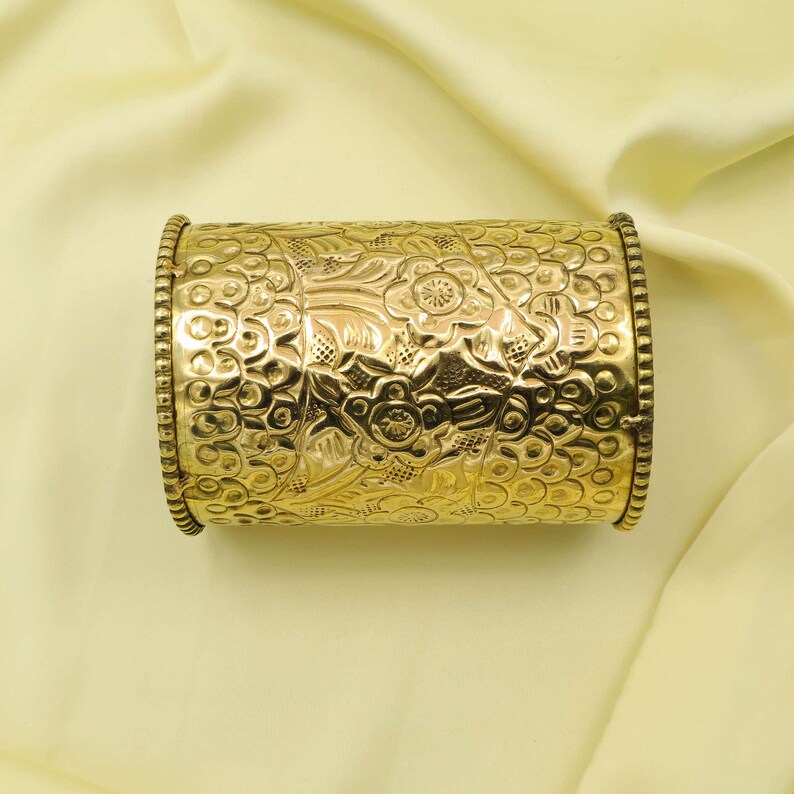May include: A gold-colored cuff bracelet with an intricate floral pattern. The bracelet has a cylindrical shape with detailed embossed designs and beaded edges. The background is a light yellow fabric.