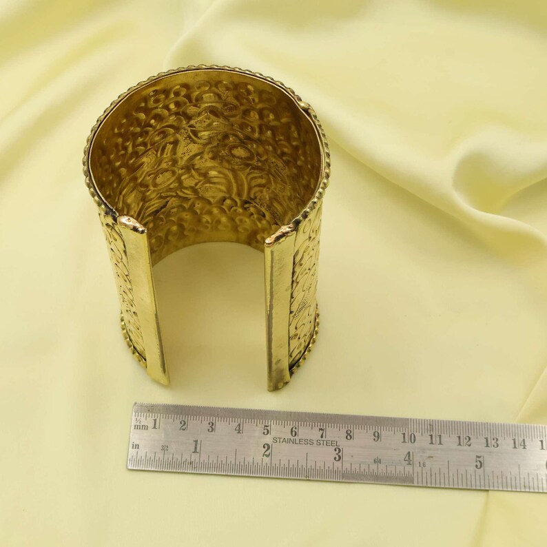 May include: A gold-colored cuff bracelet with an open design. The bracelet has an embossed floral pattern and measures approximately 1.5 inches wide. The cuff is shown on a light yellow background.