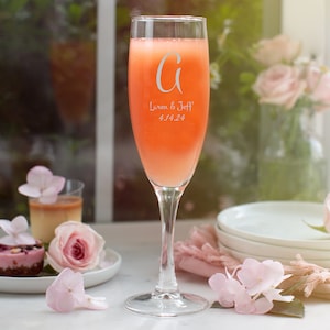 May include: A champagne flute filled with an orange-pink beverage. The glass is etched with the letter "G", the names "Lauren & Jeff", and the date "4.14.24". The scene includes desserts, flowers, and a stack of white plates, suggesting a celebration.