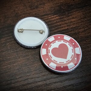 以下が含まれることがあります： 丸いピンバッジが2つ。1つは銀色のピンが付いた白いバッジ。もう1つは白いハートと白いカジノチップのデザインが描かれた赤いバッジです。
