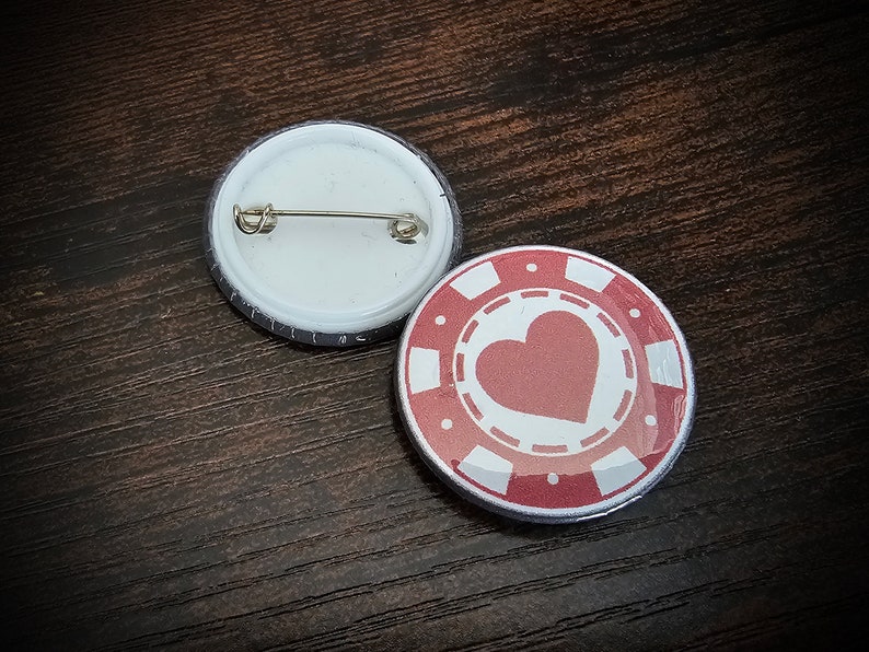 以下が含まれることがあります： 丸いピンバッジが2つ。1つは銀色のピンが付いた白いバッジ。もう1つは白いハートと白いカジノチップのデザインが描かれた赤いバッジです。
