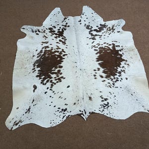 K&ouml;nnte beinhalten: Ein Kuhfellteppich mit wei&szlig;er Basis und unregelm&auml;&szlig;igen braunen Flecken. Der Teppich hat eine nat&uuml;rliche Form und Textur mit einem weichen, einladenden Aussehen. Das Fell liegt auf einer braunen Oberfl&auml;che.