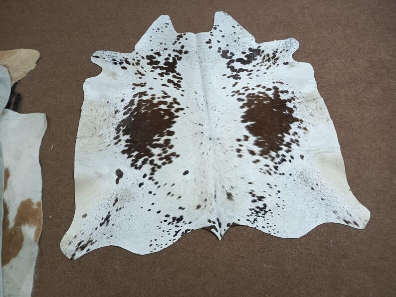 K&ouml;nnte beinhalten: Ein Kuhfellteppich mit wei&szlig;er Basis und unregelm&auml;&szlig;igen braunen Flecken. Der Teppich hat eine nat&uuml;rliche Form und Textur mit einem weichen, einladenden Aussehen. Das Fell liegt auf einer braunen Oberfl&auml;che.