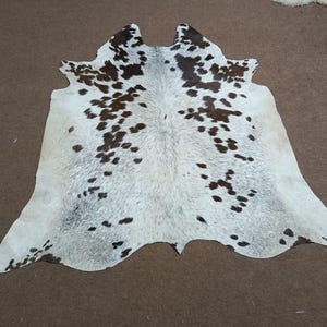 K&ouml;nnte beinhalten: Ein Kuhfellteppich mit einem nat&uuml;rlichen Muster. Der Teppich ist &uuml;berwiegend wei&szlig; mit unregelm&auml;&szlig;igen braunen Flecken. Das Fell hat eine einzigartige Form mit abgerundeten Kanten und einer Spaltung oben. Der Teppich liegt auf einer braunen Oberfl&auml;che.