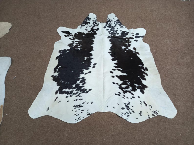 K&ouml;nnte beinhalten: Kuhfellteppich mit einem nat&uuml;rlichen Muster aus schwarzen Flecken auf wei&szlig;em Grund. Der Teppich hat eine unregelm&auml;&szlig;ige Form und eine glatte, weiche Textur. Das Kuhfell liegt auf einer braunen Oberfl&auml;che.
