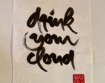 May include: A calligraphy art print on textured paper. The black ink script reads "drink your cloud." A red square stamp is in the lower right corner. The paper has a subtle, off-white color.