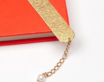 Könnte beinhalten: Ein goldenes Lesezeichen mit einer strukturierten Oberfläche und einem Perlenanhänger an einer Kette. Das Lesezeichen liegt auf einem roten Buch.