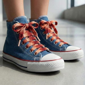 May include: Blue denim high-top sneakers with orange and red gradient laces. The sneakers have white rubber soles with a red stripe and a white toe cap. The shoes are laced up and tied in bows.