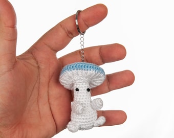 May include: White crocheted mushroom keychain with a blue top and black eyes. The keychain is attached to a silver chain.