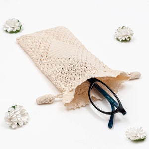 May include: A crocheted eyeglass case with a white lace design and a blue-tinted pair of reading glasses inside. The case has a tassel on the side.