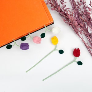 Op de afbeelding: Vijf gehaakte tulpen-bladwijzers in verschillende kleuren, waaronder paars, roze, geel, rood en wit. De bladwijzers zijn bevestigd aan een boek met een feloranje omslag.