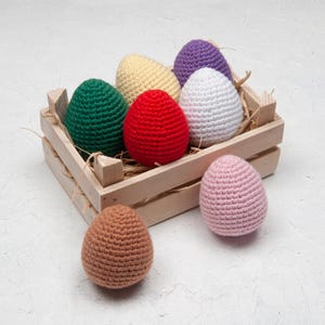 May include: A wooden crate containing a collection of colourful, crocheted Easter eggs. The eggs are in shades of green, yellow, red, white, purple, pink, and brown. The crate rests on a white surface, with some eggs outside.