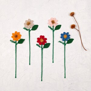 Op de afbeelding: Vijf gehaakte bloemstelen met groene bladeren en verschillende gekleurde bloemen. De bloemen zijn oranje, roze, rood, blauw en beige.