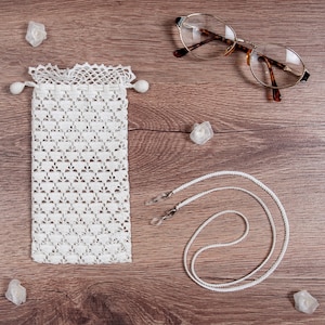 Op de afbeelding: Witte gehaakte brilhoes met een trekkoord, witte brillenketting en schildpadbril op een houten oppervlak.