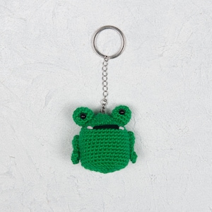 May include: Green crocheted frog keychain with black eyes. The frog is attached to a silver key ring.