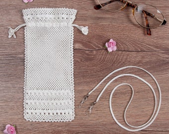 May include: White crocheted eyeglass case with a drawstring closure, a white eyeglass chain with clear clasps, and pink artificial flowers on a wood background.