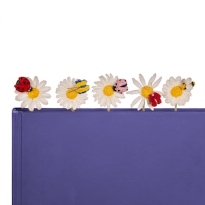 May include: Five crocheted daisy bookmarks with different insect accents. The bookmarks are white with yellow centres and are attached to a purple book.