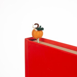 May include: A crocheted pumpkin bookmark in orange with green leaves, placed on top of a red book.