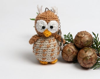 May include: A crocheted owl keychain with brown and white yarn, large white eyes, and a yellow beak. The owl is attached to a silver key ring and is sitting next to a group of brown pine cones.
