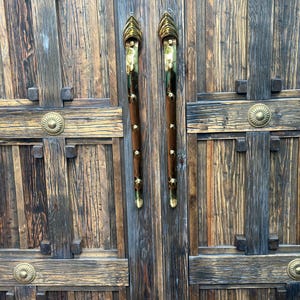 May include: Close-up of a rustic wooden door with ornate brass hardware. The door features vertical planks, decorative crossbeams, and brass handles. The handles are long, with a brown wooden grip and brass accents.