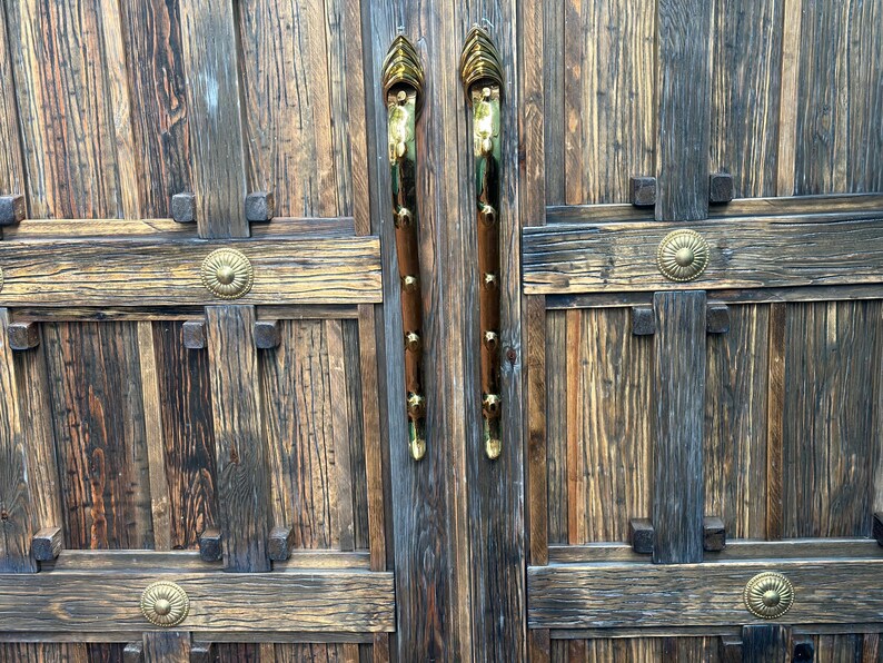 May include: Close-up of a rustic wooden door with ornate brass hardware. The door features vertical planks, decorative crossbeams, and brass handles. The handles are long, with a brown wooden grip and brass accents.