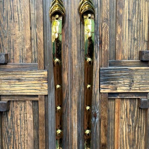 Könnte beinhalten: Zwei verzierte Türgriffe mit goldener und brauner Oberfläche. Die Griffe sind vertikal ausgerichtet und weisen eine Reihe goldener Akzente auf. Die Tür, an der sie befestigt sind, besteht aus verwittertem Holz.