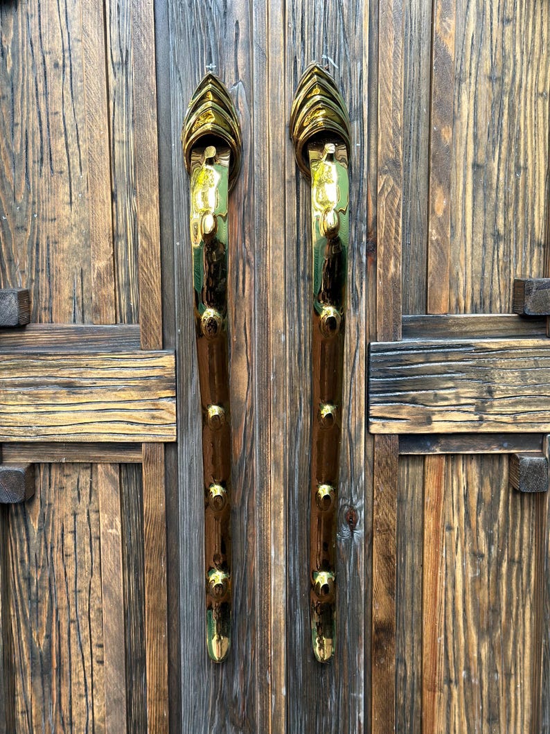 May include: Two ornate door handles with a gold and brown finish. The handles are vertically oriented and feature a series of gold accents along their length. The door they are attached to is made of weathered wood.