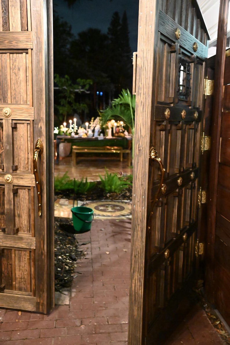 May include: A view through an open wooden gate and door. The gate and door are made of dark wood with decorative metal hardware. A brick pathway leads to a table set with candles and flowers. A green bucket sits on the pathway.