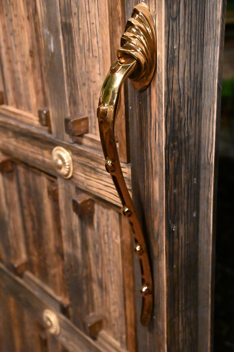 May include: A brass door handle with a curved design, mounted on a weathered wooden door. The handle has a decorative, flowing shape with small, round accents. The door is made of dark, textured wood.
