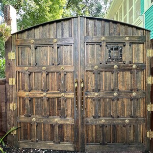 May include: A pair of large, wooden garden gates with a rustic, aged appearance. The gates are constructed from vertical planks and feature decorative metal hardware, including hinges, handles, and circular accents. A small, square window is set into the right gate.