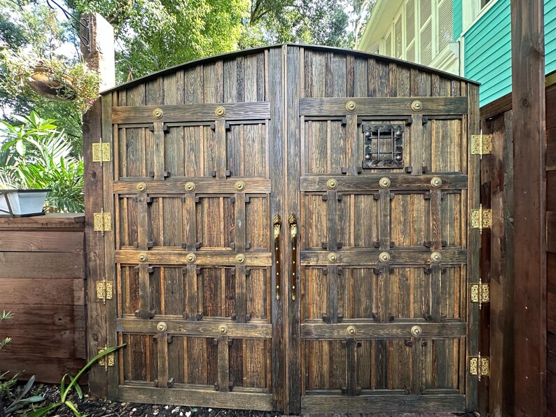 May include: A pair of large, wooden garden gates with a rustic, aged appearance. The gates are constructed from vertical planks and feature decorative metal hardware, including hinges, handles, and circular accents. A small, square window is set into the right gate.