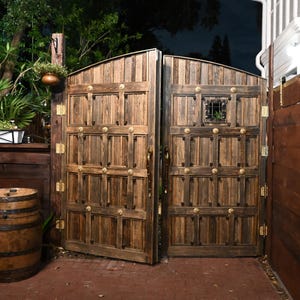 May include: A pair of large, arched wooden gates with decorative brass hardware. The gates are open, revealing a glimpse of a lush garden. A wooden barrel sits in front of the gates, and a hanging planter is visible.