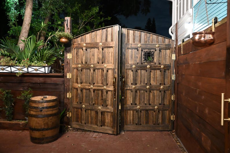 May include: A pair of large, arched wooden gates with decorative brass hardware. The gates are open, revealing a glimpse of a lush garden. A wooden barrel sits in front of the gates, and a hanging planter is visible.