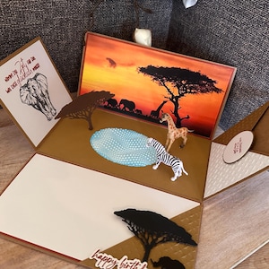 Könnte beinhalten: Eine handgefertigte Pop-up-Karte mit afrikanischem Safari-Thema. Die Karte zeigt eine Sonnenuntergangsszene, eine Giraffe, ein Zebra und einen Elefanten. Die Karte ist braun und weiß mit den Worten "happy birthday" auf der Vorderseite.