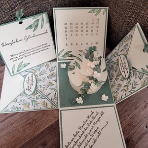 Könnte beinhalten: Eine weiße und grüne Papierbox mit floralem Design. Die Box ist geöffnet und enthält einen weißen Kuchen. Die Box hat eine Karte auf der Vorderseite, auf der "Herzlichen Glückwunsch" steht, und eine Karte im Inneren, auf der "Seite an Seite, Hand in Hand, verbunden durch der Liebe Band, wird die Sehnsucht zweier Herzen still mit den Worten Ja, ich will!" steht.