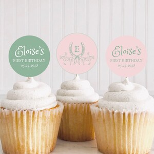 Puede incluir: Tres cupcakes con glaseado blanco cubiertos con adornos redondos de papel para cupcakes. Los adornos son rosas y verdes con el texto "Eloise's First Birthday 05.25.2026" y un diseño de herradura.