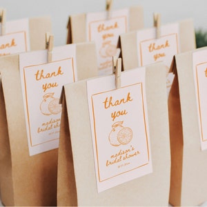 Peut inclure: Sacs cadeaux en papier brun avec des étiquettes blanches portant l'inscription "merci" en orange. L'étiquette présente une illustration d'orange et le texte "madison's bridal shower". Chaque sac est fixé avec une petite pince à linge en bois.
