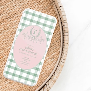 Puede incluir: Funda de teléfono con estampado de cuadros vichy verdes y blancos. Una superposición ovalada rosa muestra el texto "Eloise's First Birthday" e ilustraciones ecuestres. La funda está sobre una bandeja de mimbre trenzado.