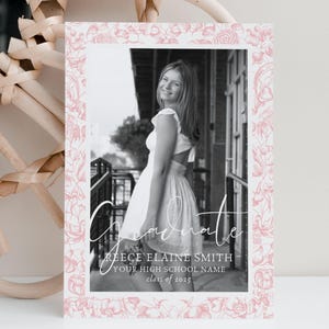 May include: A graduation announcement featuring a black and white photo of a person in a white dress. The photo is framed with a pink floral border. The word "Graduate" is written in script, along with the graduate's name, high school name, and class year.