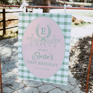 Puede incluir: Un cartel de bienvenida de cuadros vichy verde y blanco con un óvalo rosa en el centro. El óvalo tiene una herradura verde con la letra "E" en el centro. El texto "Bienvenido a Eloise's First Birthday" está escrito en verde sobre el óvalo rosa.