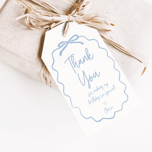 Op de afbeelding: Een cadeau verpakt in beige papier en vastgebonden met raffia. Een wit label met een blauwe geschulpte rand en strik leest "Thank You" met een verjaardagsbericht en een naam.