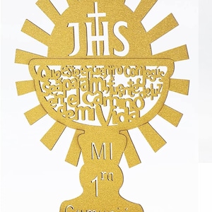 May include: Gold glitter first communion centerpiece with a chalice design and the words "JHS" and "MI 1ra Comunión".