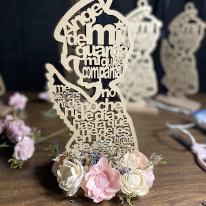 May include: A wooden angel figurine with the words "Angel de mi guarda, mi dulce compania" and other Spanish text. The figurine is decorated with a small bouquet of pink and white flowers.