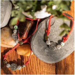 May include: Two red string bracelets with silver charms. The charms feature religious imagery and are surrounded by small silver beads. The bracelets are lying on a wooden surface with stones.