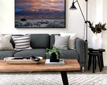 May include: A large framed landscape photograph of a mountain range at sunset with golden clouds hanging over the peaks, displayed on a white wall above a dark gray sofa, a wooden coffee table, and a black wall-mounted lamp with a white vase.
