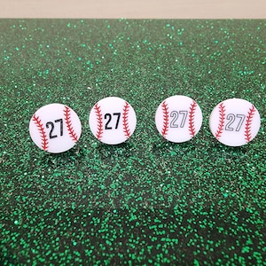 May include: Four white baseball-themed ornaments with red stitching and the number 27 printed in black. The ornaments are on a green glitter background.
