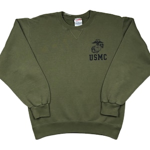 May include: Olive green sweatshirt with the USMC emblem printed on the front. The crew neck sweatshirt has long sleeves and ribbed cuffs and hem. The text "USMC" is printed below the emblem.